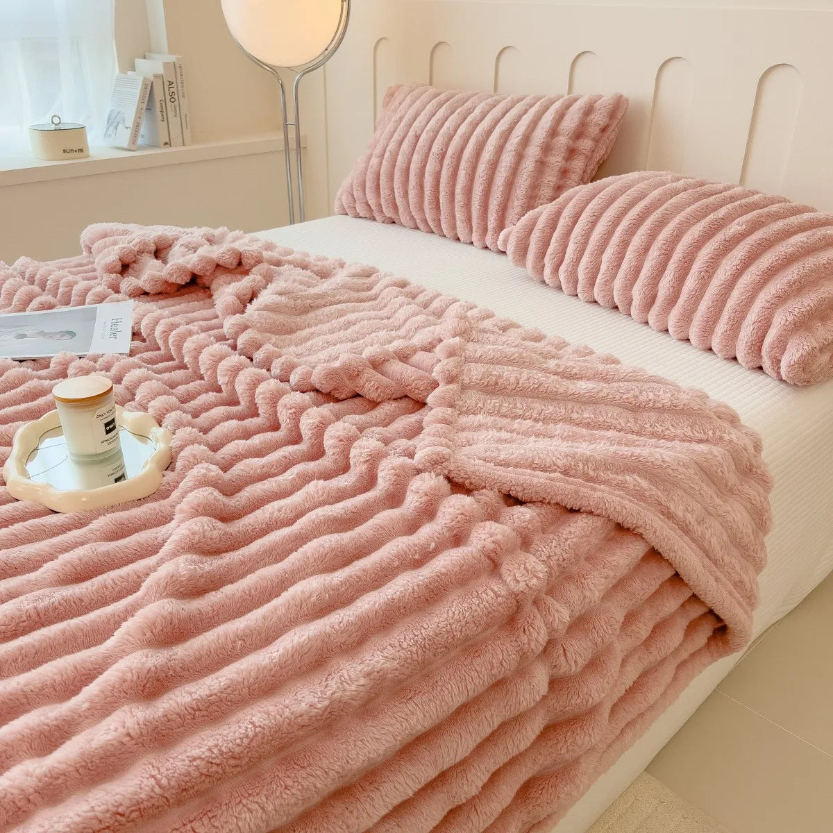 A thick pink fuzzy blanket is draped over a white bed, with a matching striped throw rolled up beside it.