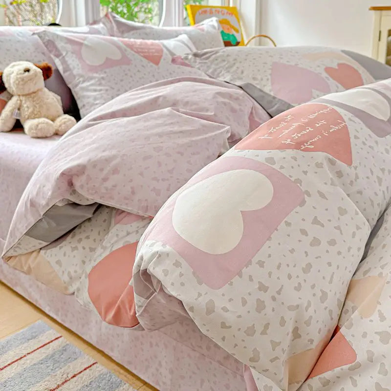 A pink and gray duvet and pillow set featuring heart patterns and a teddy bear plush toy.