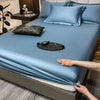 A light blue fitted bed sheet is draped over a bed, with several matching pillows and a black leather item on the surface.
