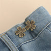 A pair of gold-toned clover button clips are fastened onto the fabric of a denim garment.