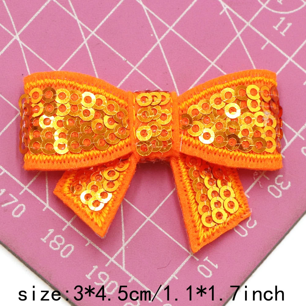 Orange sequin bow patches for DIY decoration shown on a pink grid background