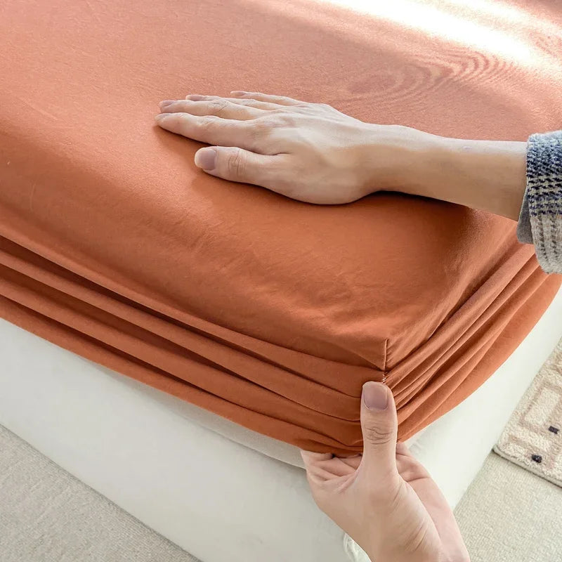 A hand is folding a terracotta orange fitted bed sheet on a white surface.