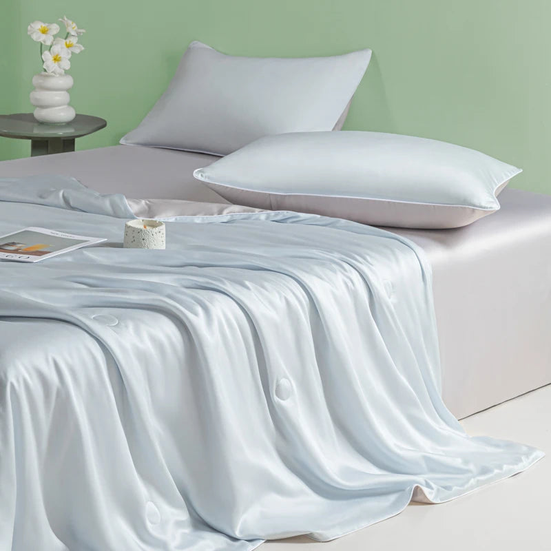 A light blue ice silk cooling quilt draped over a bed with white pillows and a pale green wall.