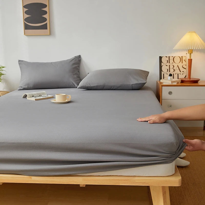 A gray fitted sheet is laid on a wooden bed, with pillows and a lamp visible in the background.
