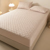A white quilted mattress protector is shown on a bed, next to a lamp.