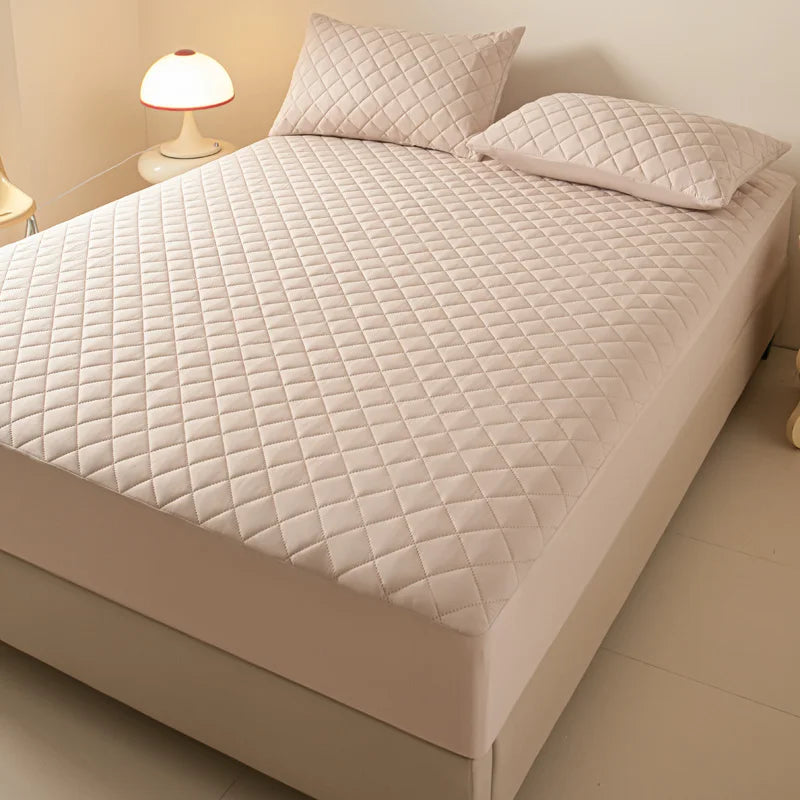 A white quilted mattress protector is shown on a bed, next to a lamp.