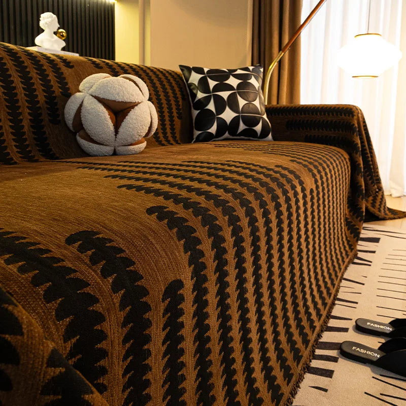 A close-up of a brown herringbone chenille sofa cover with black geometric patterns and a white tassel accent pillow.