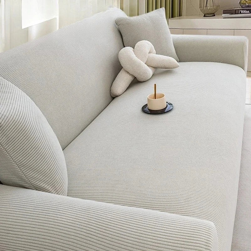 A light beige stretch jacquard sofa slipcover in a living room setting with a decorative knitted pillow.