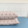 A set of two plaid beige and brown pillow covers resting on a blue surface against a white wainscoting background.