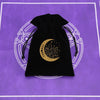 A black velvet pouch with a gold crescent moon and star design rests on a purple mat.