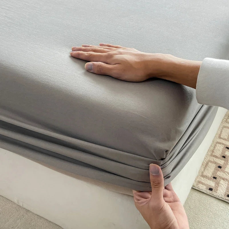 A hand is adjusting a gray fitted bed sheet on a mattress.