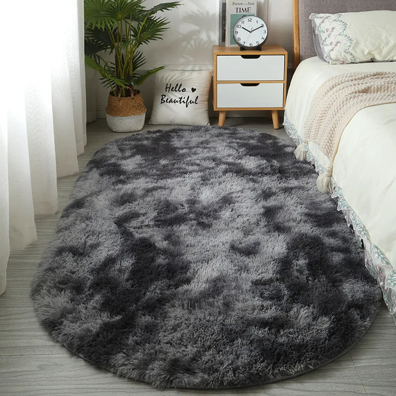 A dark gray plush floor mat placed on a light wood floor next to a white bed and a wooden nightstand.