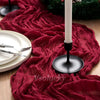 A deep red crinkled cheesecloth table runner draped across a wooden surface with a white candle and greenery.