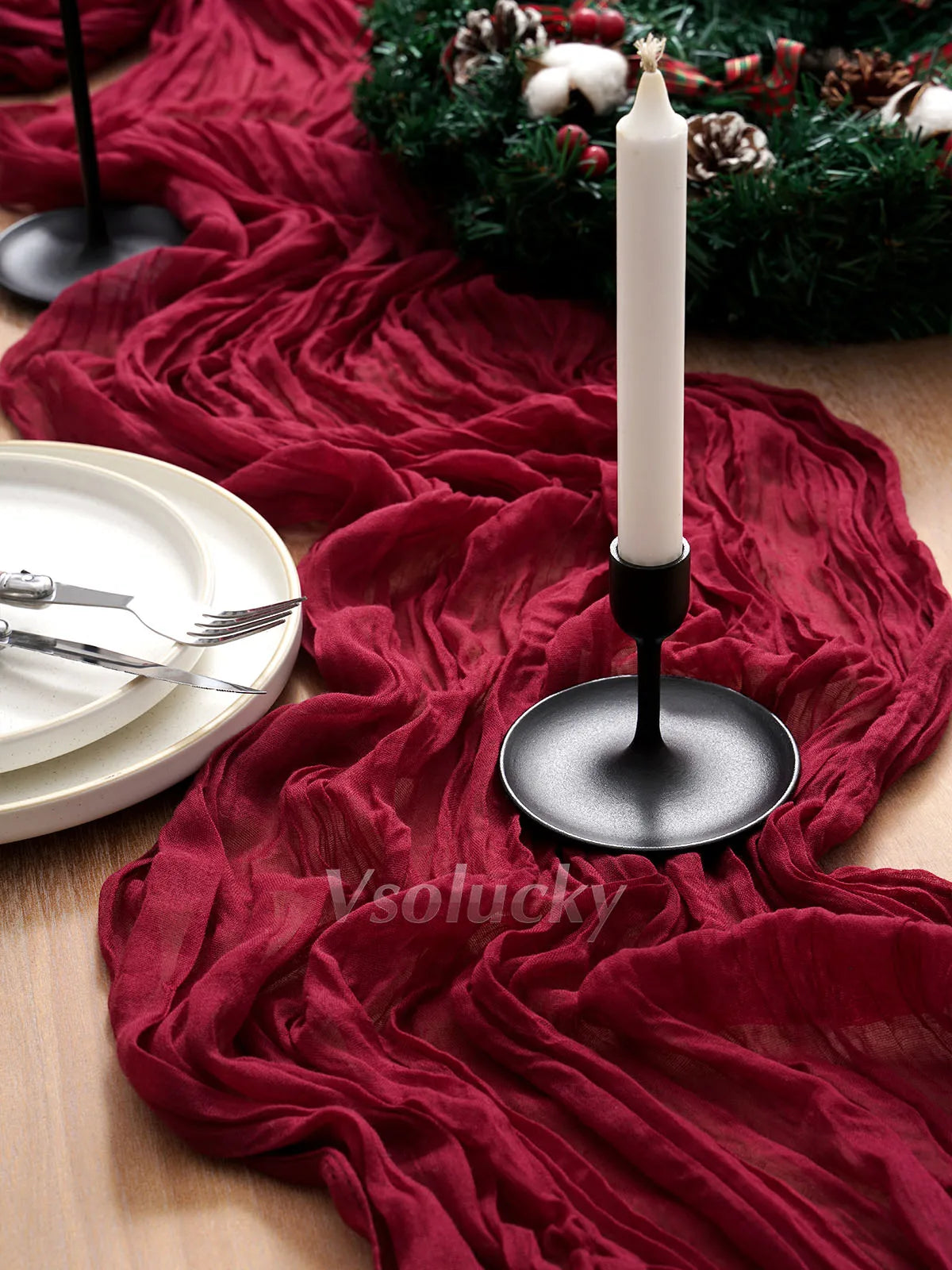 A deep red crinkled cheesecloth table runner draped across a wooden surface with a white candle and greenery.