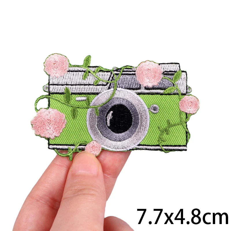 Embroidered iron-on patch featuring a stylized camera design with pink floral accents on a green background