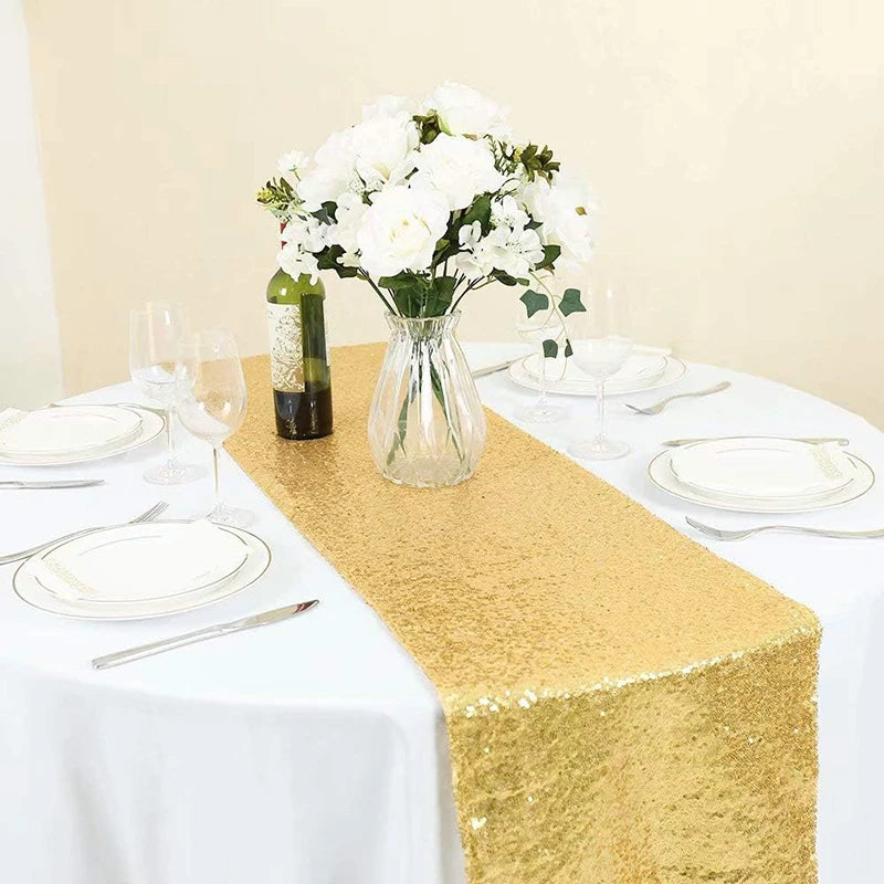 A golden sequin table runner on a white tablecloth with white floral arrangements and place settings.