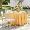 A round table set with a golden tablecloth, white plates, silverware, and a floral centerpiece outdoors.