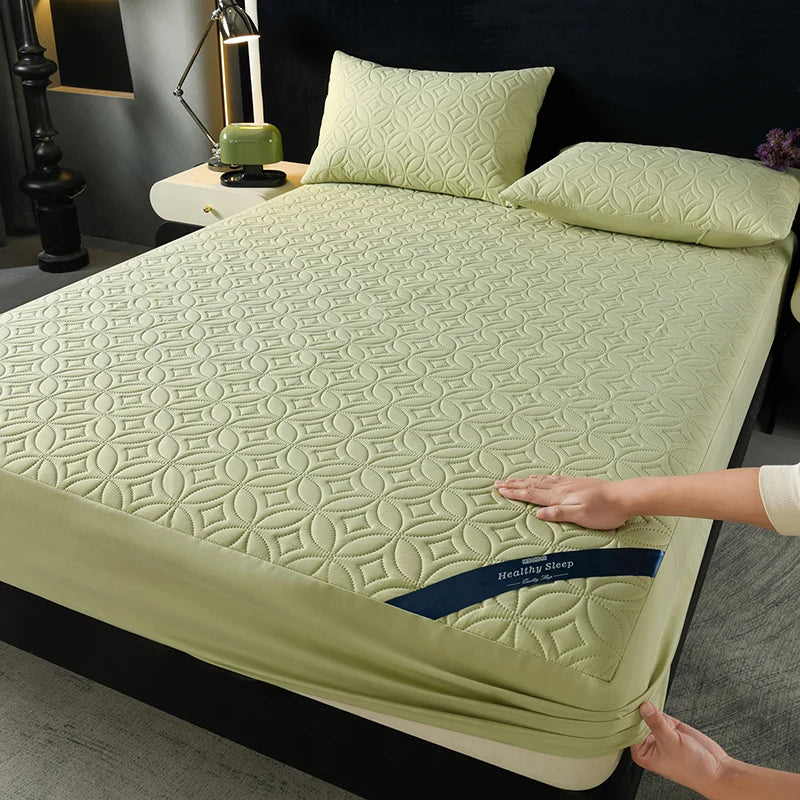 A light green quilted mattress protector is shown on a bed, with a hand touching the fabric.