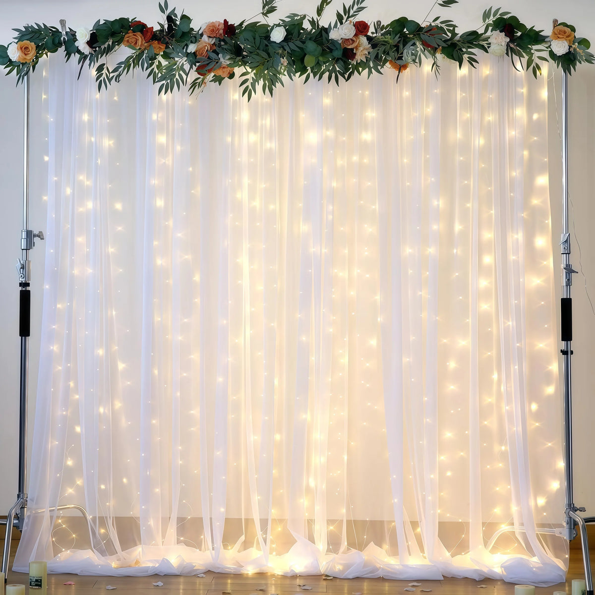 A sheer white tulle backdrop curtain decorated with warm fairy lights and floral greenery for an event.