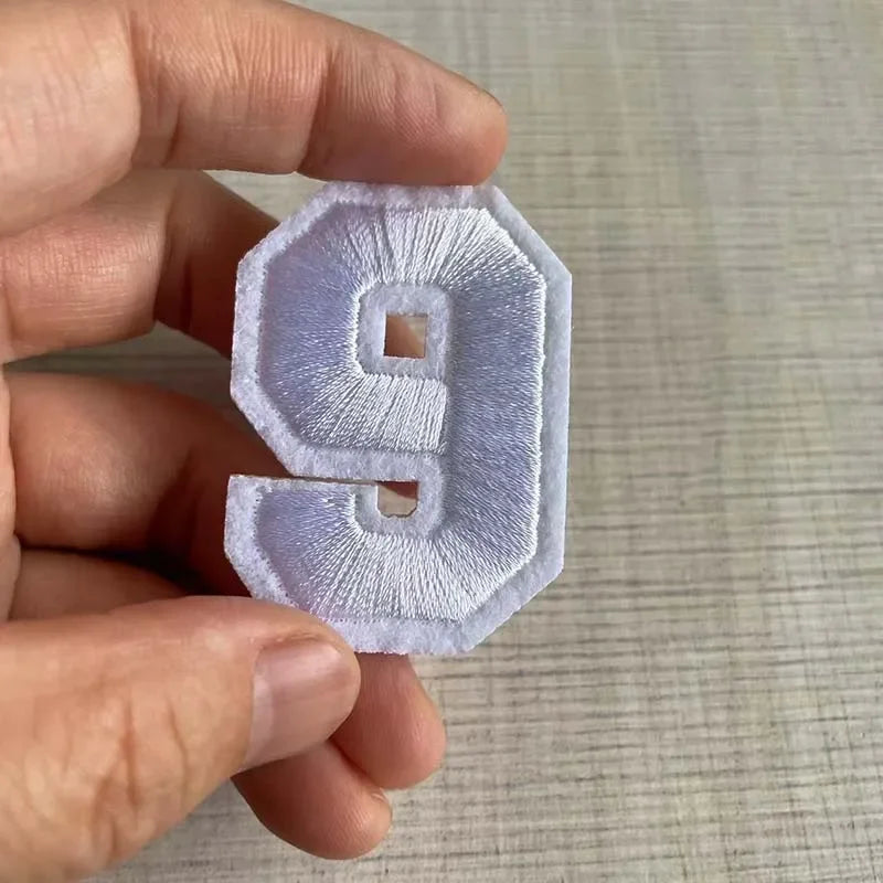 A hand holds a white embroidered number 9 patch for apparel.