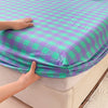 A hand is touching a purple and green checkered fitted sheet on a bed.