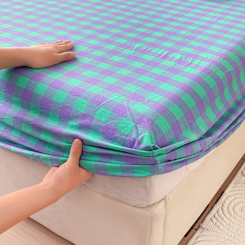 A hand is touching a purple and green checkered fitted sheet on a bed.