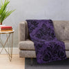 A dark purple gothic damask throw blanket draped over a gray sofa on a light rug.