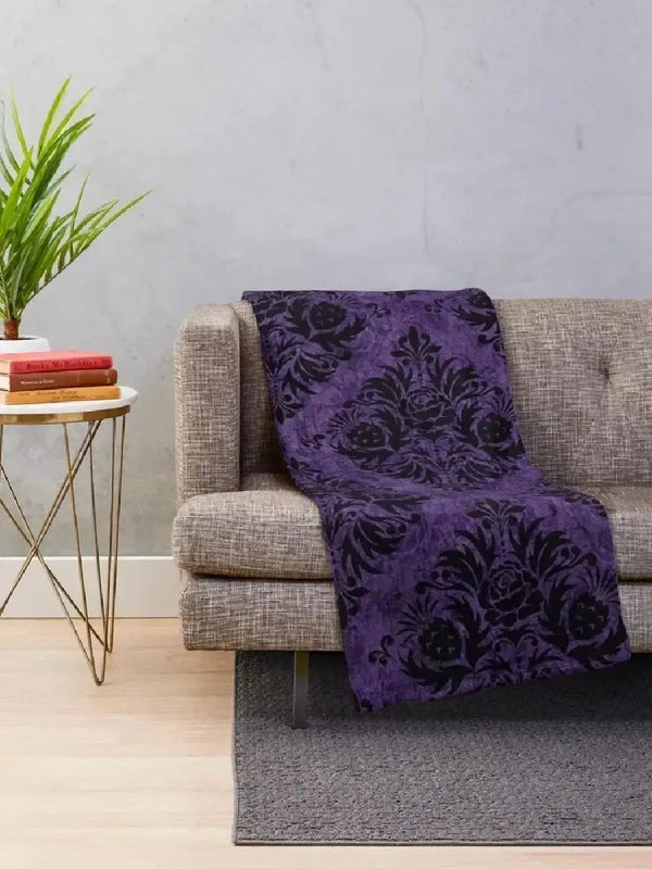A dark purple gothic damask throw blanket draped over a gray sofa on a light rug.