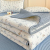 A stack of white and blue floral quilted bedspreads and a gray quilted pillow on a light blue bed.