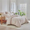 A beige luxury quilt duvet set displayed on a bed in a bright, minimalist room with white window blinds and green plants.