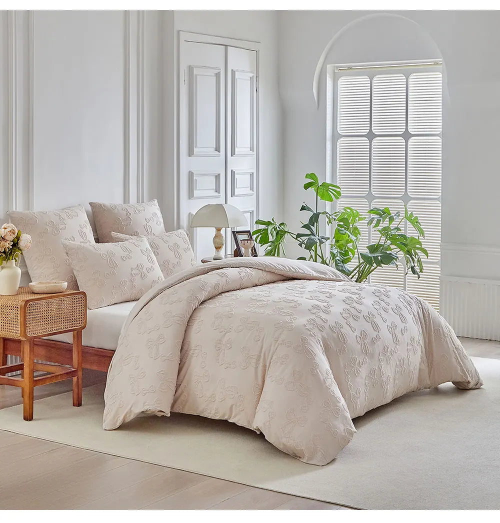 A beige luxury quilt duvet set displayed on a bed in a bright, minimalist room with white window blinds and green plants.