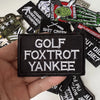 A hand holds a black iron-on slogan patch featuring the text GOLF FOXTROT YANKEE among other patches.