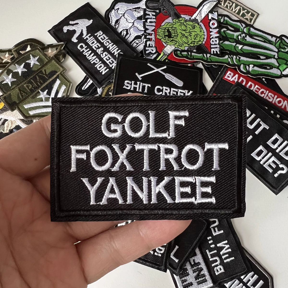 A hand holds a black iron-on slogan patch featuring the text GOLF FOXTROT YANKEE among other patches.