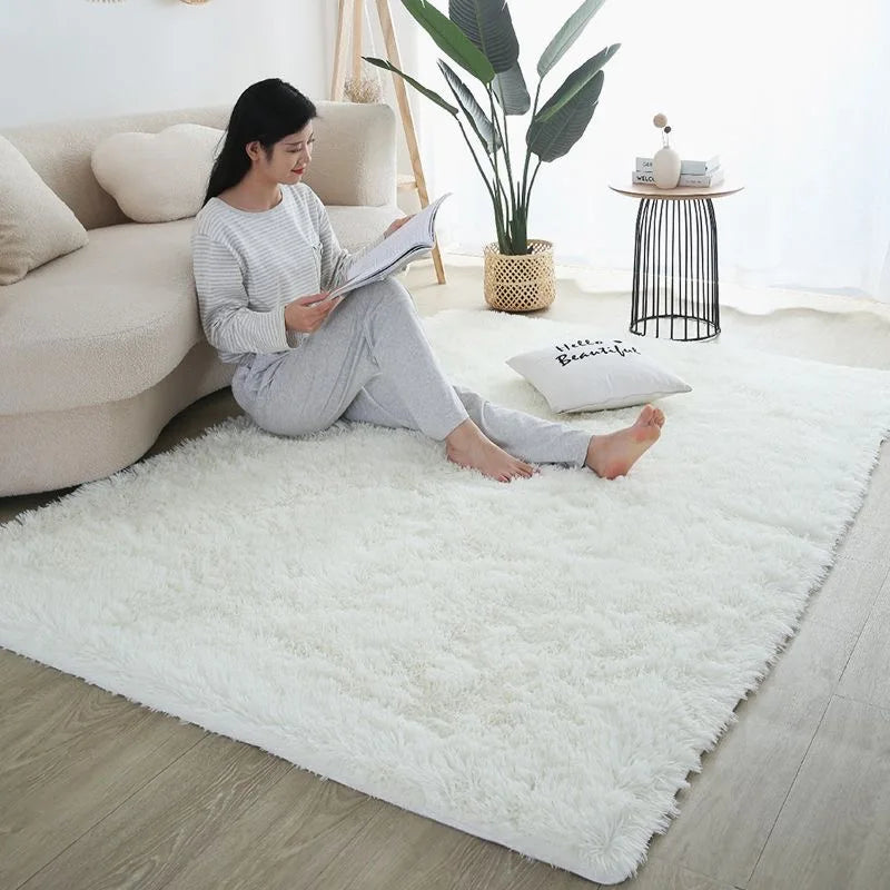 A woman sits on a white fluffy rug reading a book in a bright living room with indoor plants and minimalist decor.