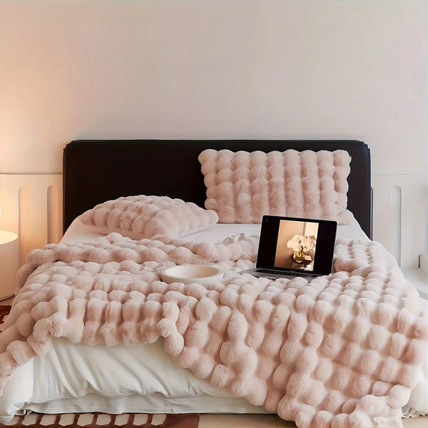 A fluffy pink faux rabbit fur throw blanket draped over a white bed with a matching pillow and dark headboard.