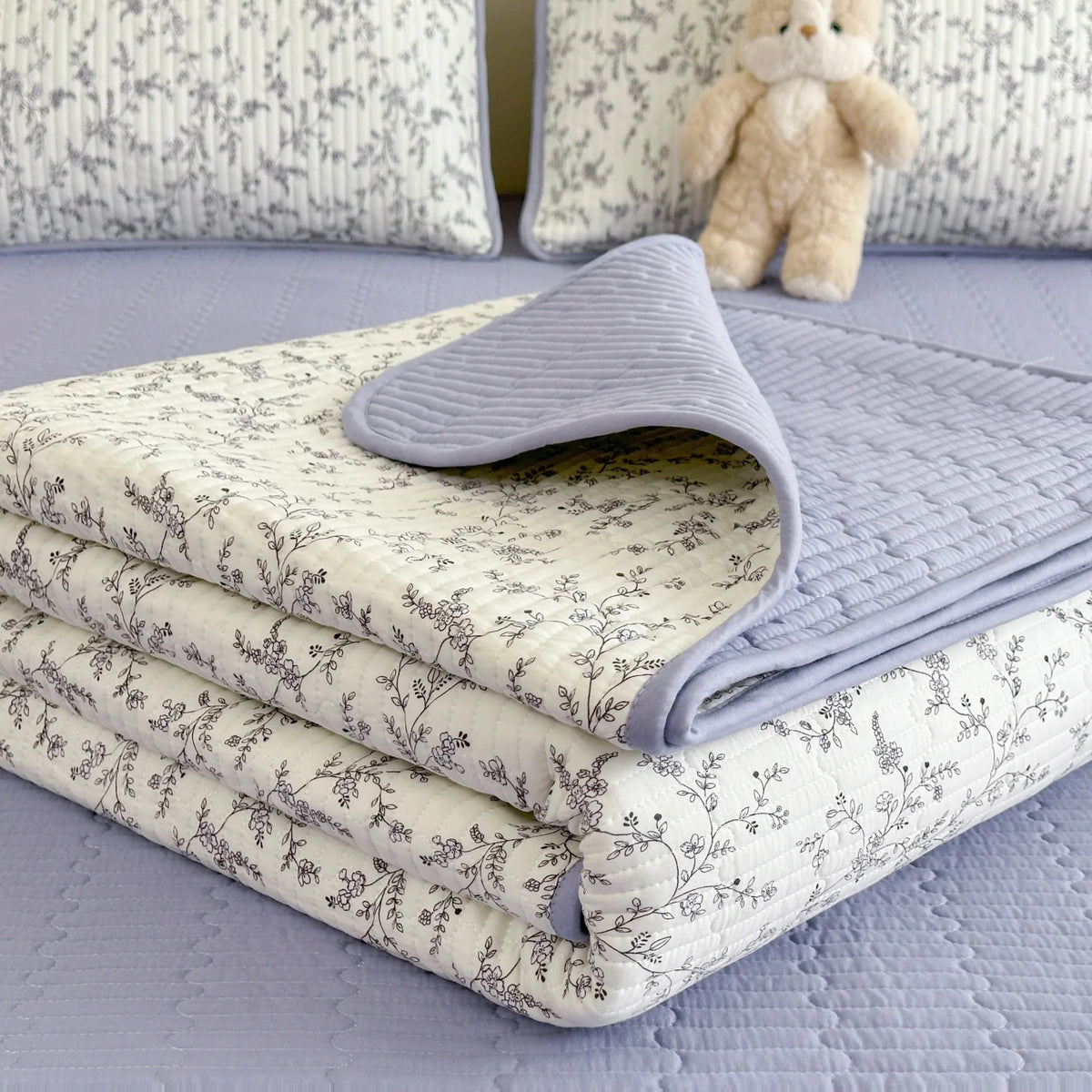 A stack of luxury floral quilted bedspreads in white and gray tones resting on a lavender surface.