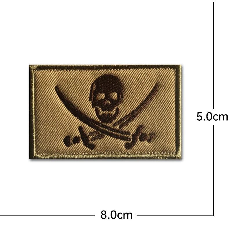 A rectangular embroidery patch featuring a black pirate skull and crossed swords on a tan fabric background.