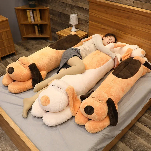A woman is relaxing on a gray surface surrounded by several large, soft orange and beige dog plush pillows.