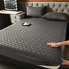 A dark gray quilted mattress protector is shown on a bed, with pillows and a tray nearby.