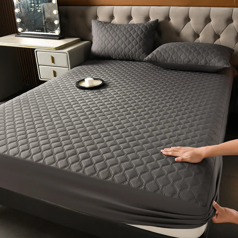A dark gray quilted mattress protector is shown on a bed, with pillows and a tray nearby.