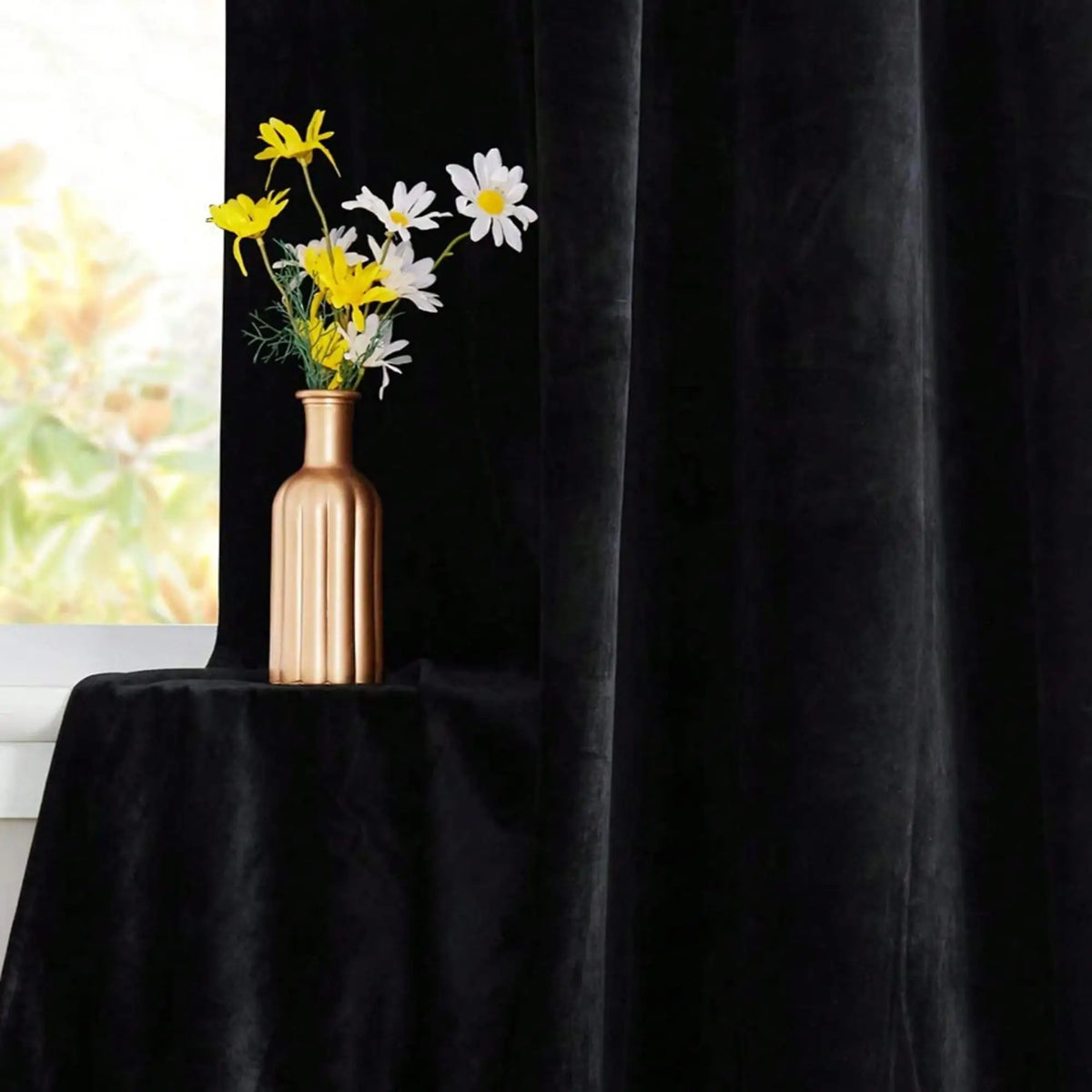 A vase of yellow and white flowers sits on a black velvet draped table next to a black curtain.