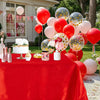 A red satin tablecloth is set for an elegant outdoor party with pink and gold balloons, flowers, and desserts.