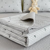 A stack of light gray quilted fabric and pillows with a small yellow stuffed rabbit on a bed.