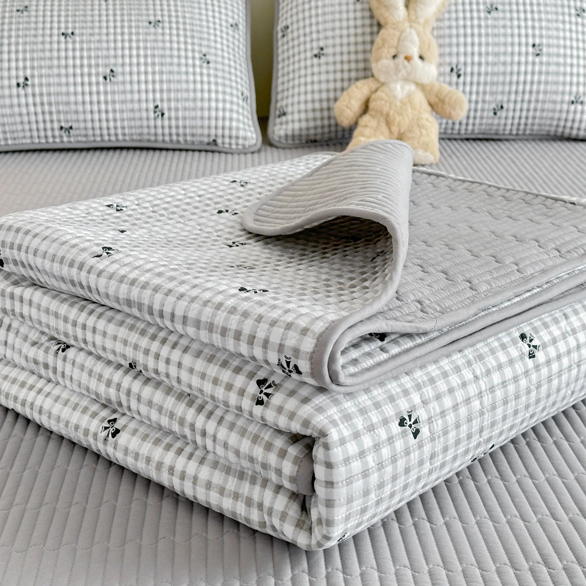A stack of light gray quilted fabric and pillows with a small yellow stuffed rabbit on a bed.