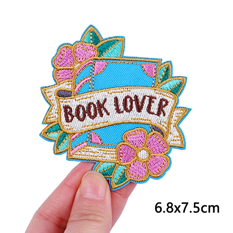 An embroidered iron-on patch featuring the text BOOK LOVER surrounded by floral designs in pink, teal, and yellow thread.