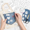 Hands are sewing a piece of denim fabric with white embroidery patterns and a yellow canvas bag in a bright studio setting.
