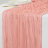 A flowing, dusty rose colored rustic gauze table runner draped elegantly on a white surface.