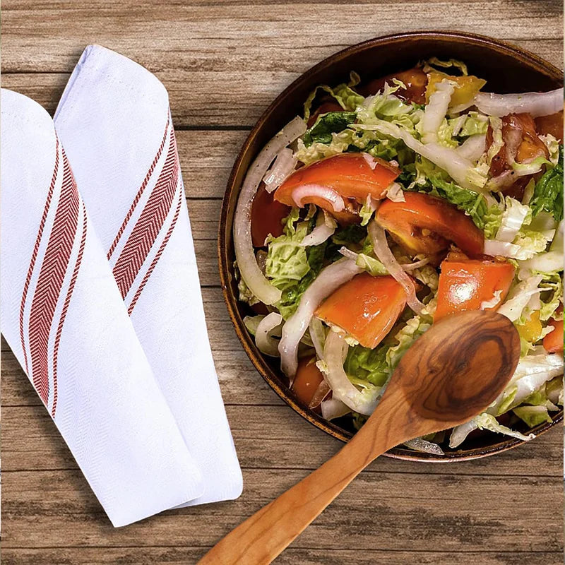 A wooden spoon rests beside a bowl of fresh salad with lettuce, carrots, and onions, next to a stack of white striped kitchen