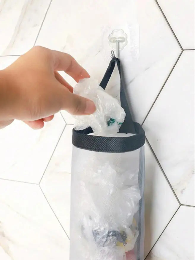 A hand is holding a white plastic bag dispenser rack with a black strap and a clear plastic container.