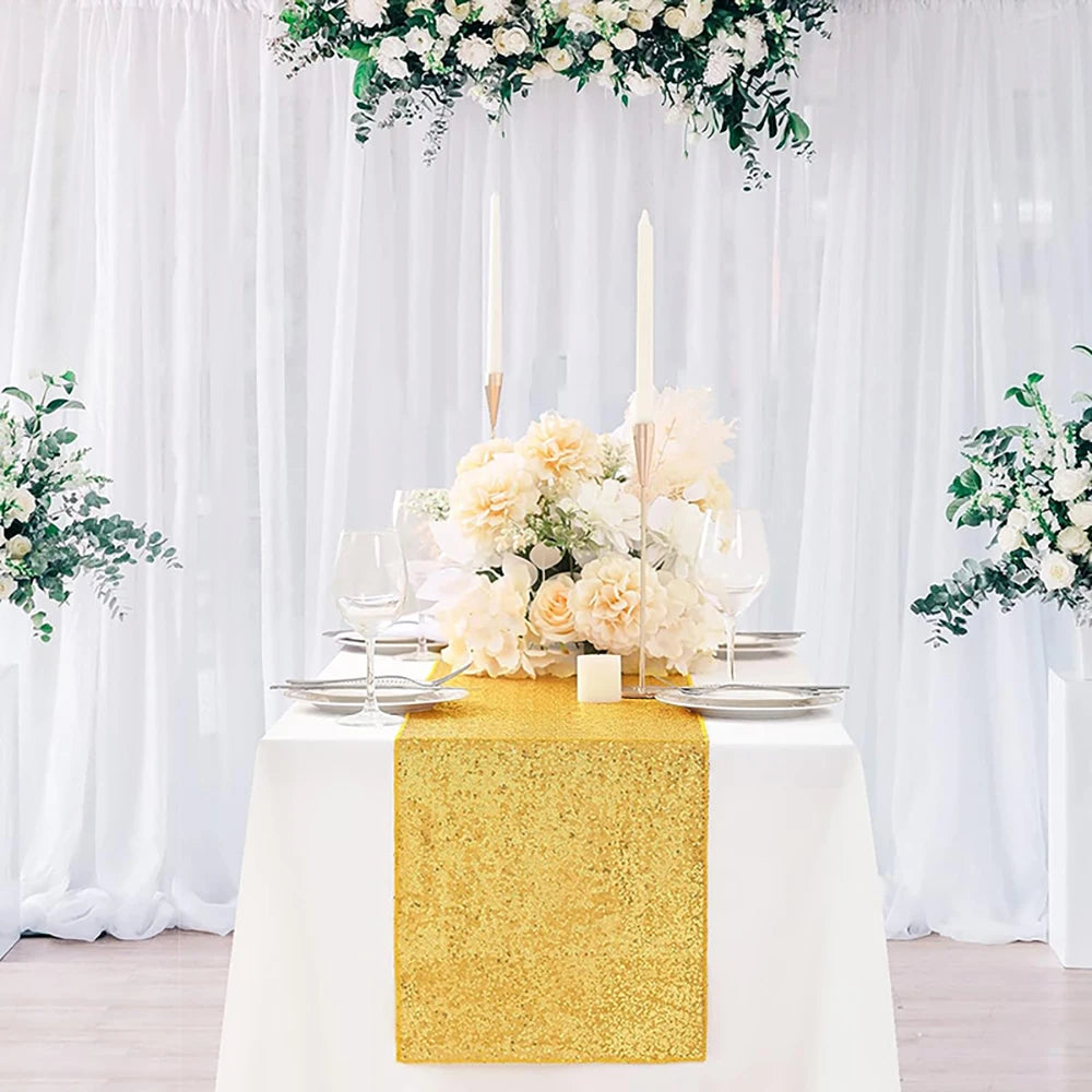 A white table with a golden sequin runner, floral centerpiece, and sheer white curtains.